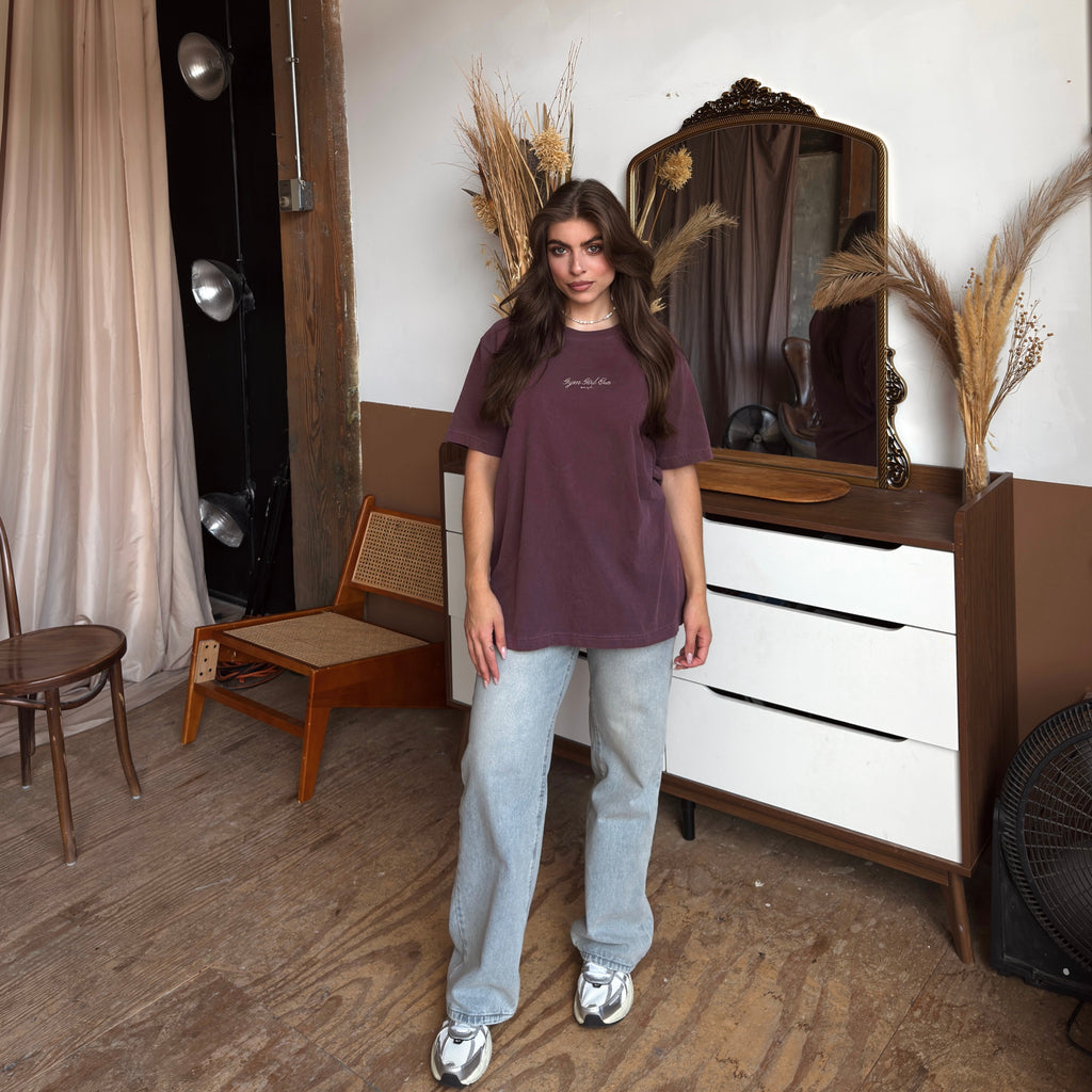 Person wearing a maroon t-shirt and light blue jeans standing in a room with a white wall, wooden floor, and decorative elements.